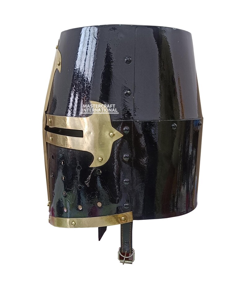 Op de afbeelding: Een zwarte, glanzende ridderhelm met messingkleurige accenten. De helm heeft een afgeronde bovenkant en een gezichtsbescherming met decoratieve messing elementen. De tekst "MASTER CRAFT INTERNATIONAL" is zichtbaar. Klinknagels bevestigen de metalen platen.