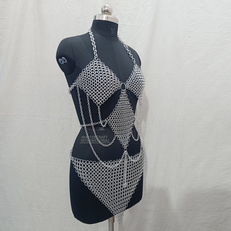 May include: A silver-toned chainmail body harness featuring a halter-style top, a diamond-shaped midsection panel, and a triangular lower section, all constructed from interlocking metal rings. The text MASTERCRAFT INTERNATIONAL is visible.