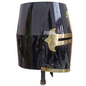 Op de afbeelding: Een zwarte, glanzende ridderhelm met messing details. De helm heeft een geklonken constructie en een messing frontplaat. De tekst "MASTERCRAFT INTERNATIONAL" is zichtbaar. Een leren riem met een gesp is bevestigd.
