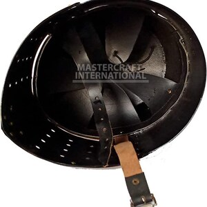 Op de afbeelding: Zwarte metalen helm met binnenvoering en kinband. De binnenkant heeft een reeks gebogen metalen platen. De helm heeft kleine ventilatiegaten rond de rand. De tekst "MASTER CRAFT INTERNATIONAL" is zichtbaar in de helm.
