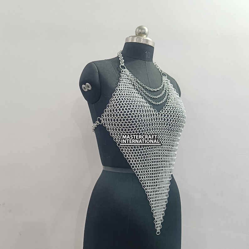 May include: Silver chainmail top with a V-neckline. The top is made of small, interlocking metal rings and has a triangular shape. The top is displayed on a black mannequin.