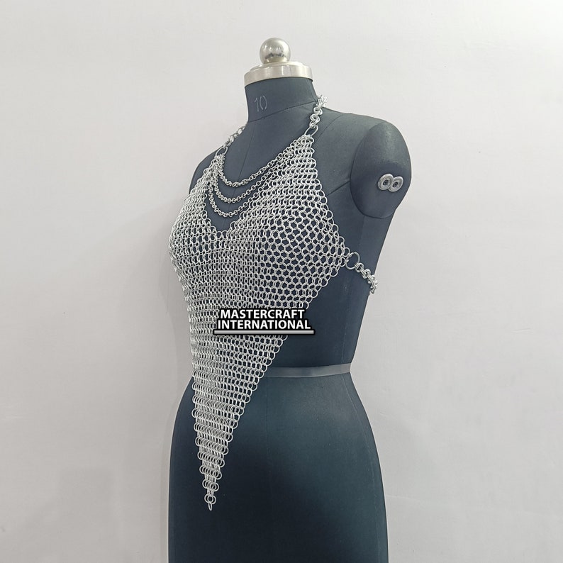 May include: Silver chainmail top with a V-neckline and a pointed hem. The top is made of small, interlocking metal rings. The text "MASTERCRAFT INTERNATIONAL" is visible on the top.