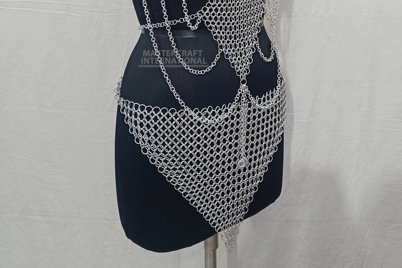May include: A silver-colored chainmail body harness featuring interlocking metal rings and draped chain accents. The piece includes a triangular front panel and is displayed on a black mannequin. The text MASTERCRAFT INTERNATIONAL is visible.