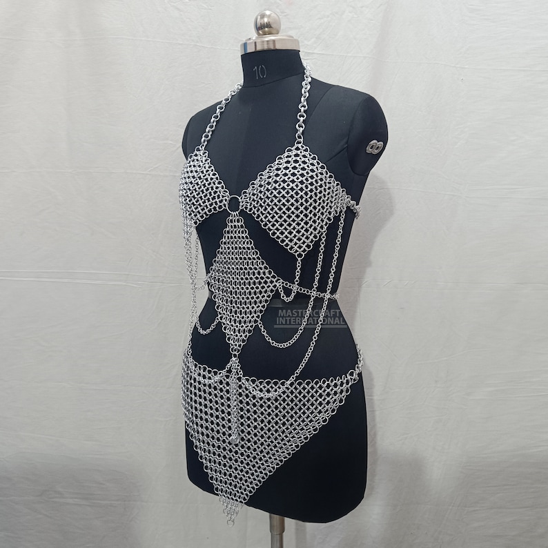 May include: A silver-colored chainmail body harness featuring a halter-style top with triangular cups, a central diamond-shaped panel, and a lower triangular skirt section. The piece is constructed from interlocking metal rings and includes the text MASTERCRAFT INTERNATIONAL printed on the midsection.