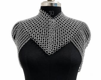 May include: A silver-toned chainmail shoulder mantle constructed from interlocking metal rings. The garment features a high collar and a pointed front design that drapes over the shoulders and upper chest, creating a textured, metallic appearance.