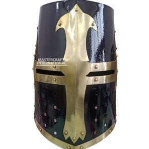 Op de afbeelding: Een zwarte middeleeuwse kruisvaarderhelm met een gouden kruisontwerp aan de voorkant. De helm heeft een glanzende afwerking en is versierd met klinknagels en gouden sierlijsten. De tekst "MASTERCRAFT INTERNATIONAL" is zichtbaar.