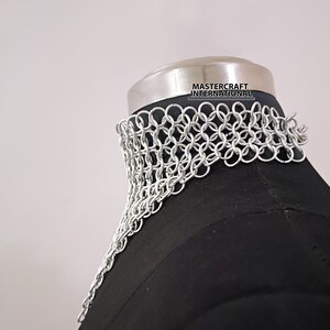 May include: Silver chainmail choker necklace on a black mannequin. The chainmail is made of small, interlocking metal rings. The necklace is labeled "Mastercraft International."