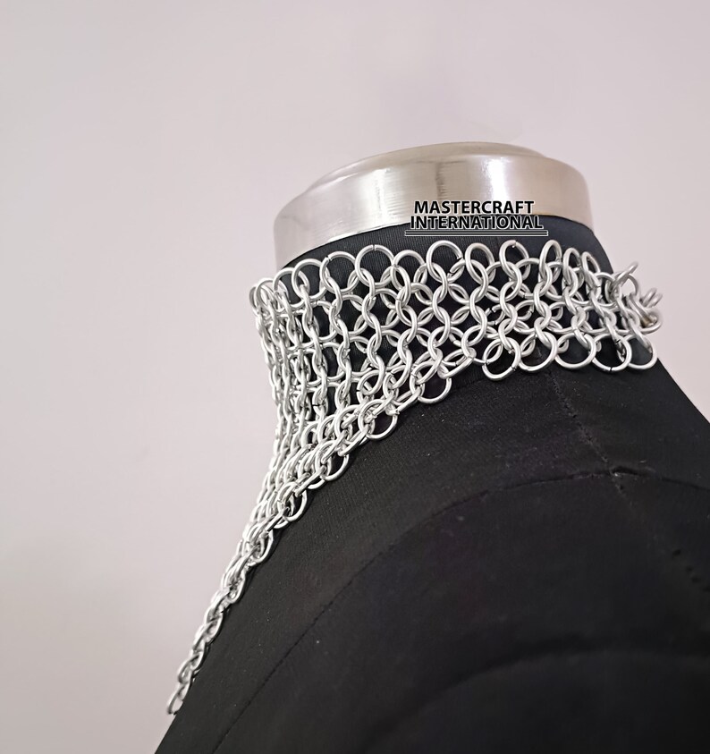 May include: Silver chainmail choker necklace on a black mannequin. The chainmail is made of small, interlocking metal rings. The necklace is labeled "Mastercraft International."