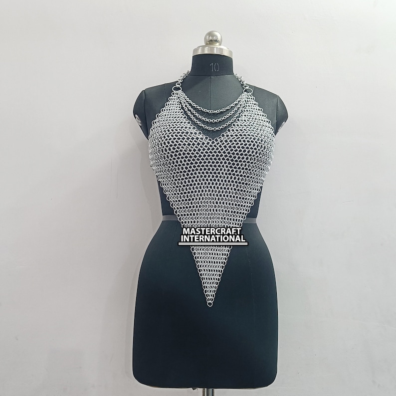 May include: A silver chainmail top with a V-neckline and a pointed bottom. The top is on a black mannequin. The text "MASTER CRAFT INTERNATIONAL" is printed on the top.