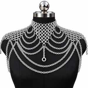 May include: A silver-toned chainmail shoulder necklace featuring a high-neck collar design. The piece is constructed from interlocking metal rings that drape across the chest and shoulders in multiple layered strands, ending in a central ring.