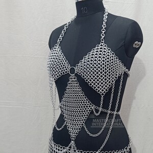 May include: A silver-colored chainmail halter top featuring a diamond-shaped bodice panel, a neck strap, and dangling chain accents. The garment is displayed on a black mannequin and includes the text MASTERCRAFT INTERNATIONAL printed on the lower right.