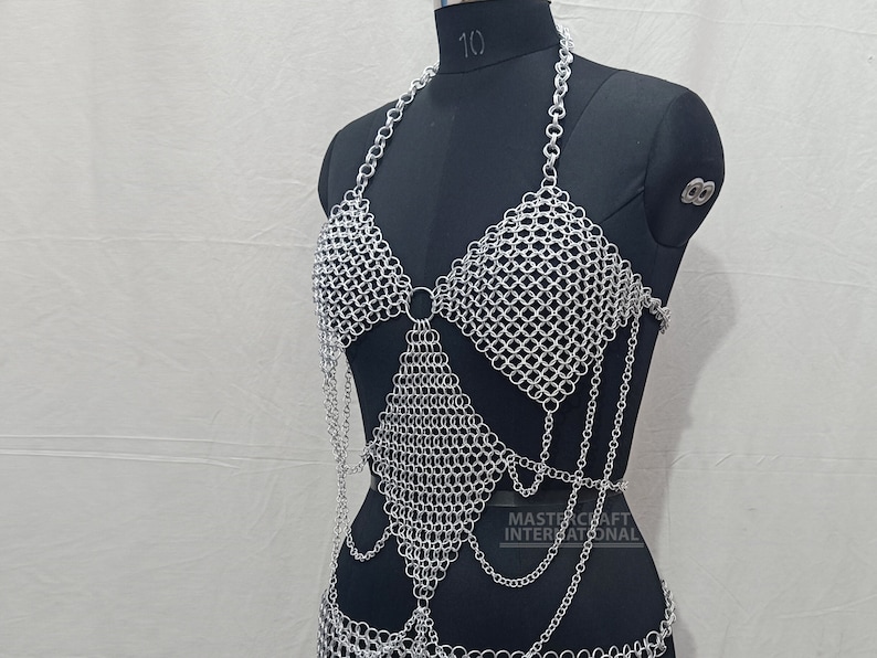 May include: A silver-colored chainmail halter top featuring a diamond-shaped bodice panel, a neck strap, and dangling chain accents. The garment is displayed on a black mannequin and includes the text MASTERCRAFT INTERNATIONAL printed on the lower right.
