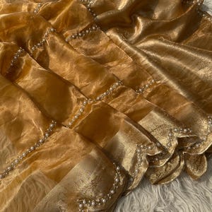 May include: Close-up of a golden fabric with a shimmering, translucent appearance. The fabric is adorned with delicate rows of small, white pearls. The edges feature a scalloped design, adding a touch of elegance to the textile.