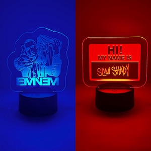 May include: Two LED night lights. One features a silhouette of a man holding a boombox with the name "EMINEM" below. The other is a red light with the text "HI! MY NAME IS SLIM SHADY". Both lights sit on black bases.