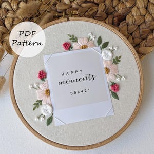 Może przedstawiać: Haftowane koło z kwiatami na kremowym tle. Projekt zawiera różowe i białe kwiaty, zielone liście i czerwone jagody. Na środku biała karta z napisem "HAPPY moments" o wymiarach 8,9 x 10,7 cm.