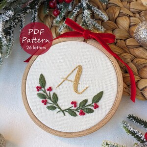 Pode incluir: Bordado em bastidor com a letra "A" em fio dourado, rodeado por uma grinalda de folhas verdes e bagas vermelhas. O bastidor de madeira tem uma fita vermelha para pendurar. O texto "PDF Pattern 26 letters" está num círculo vermelho.