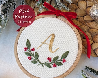 Pode incluir: Bordado em bastidor com a letra "A" em fio dourado, rodeado por uma grinalda de folhas verdes e bagas vermelhas. O bastidor de madeira tem uma fita vermelha para pendurar. O texto "PDF Pattern 26 letters" está num círculo vermelho.