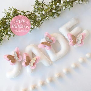 Könnte beinhalten: Weiße Filzbuchstaben mit der Aufschrift "JADE", verziert mit rosa und goldfarbenen Glitzer-Schmetterlingen. Die Buchstaben sind auf einer weißen Oberfläche angeordnet, mit einer Schnur aus weißen Pompons und floralen Akzenten. Das Bild enthält einen rosa Kreis mit dem Text "PDF Pattern 26 letters."