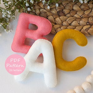 Könnte beinhalten: Drei Filzbuchstaben: ein rosa "B", ein weißes "A" und ein gelbes "C". Die Buchstaben sind mit einem Kontrastfaden handgenäht. Ein rosa Kreis mit dem Text "PDF Pattern 26 letters" befindet sich in der unteren linken Ecke. Die Buchstaben liegen auf einer weißen Oberfläche.