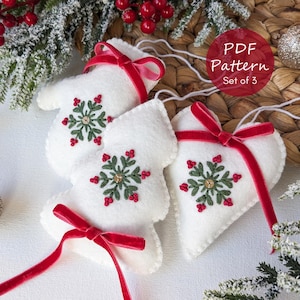Puede incluir: Conjunto de tres adornos navideños de fieltro blanco con forma de mitón, árbol y corazón. Cada adorno presenta un diseño de copo de nieve bordado en verde y rojo y una cinta de terciopelo roja. El texto "PDF Pattern Set of 3" está en un círculo rojo.