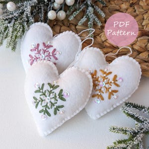 Pode incluir: Enfeites de coração de feltro branco com desenhos de flocos de neve bordados em rosa, verde e dourado, adornados com lantejoulas. Os enfeites têm uma borda ondulada e uma argola para pendurar. A imagem inclui um rótulo de padrão PDF.