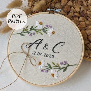 Può includere: Ricamo su telaio con le iniziali "A & C" e la data 12.07.2025. Il design presenta margherite bianche, fiori viola e foglie verdi. Il testo "PDF Pattern" è in un cerchio bianco. Il ricamo è su un tessuto beige.