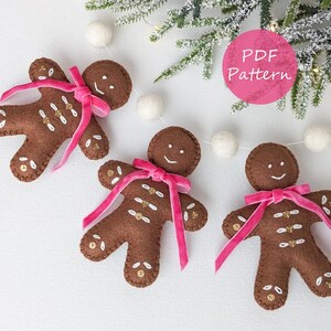 Op de afbeelding: Drie bruine vilten peperkoekmannetjes met roze strikken. Elk ornament heeft witte stiksels en gouden pailletten accenten. De ornamenten maken deel uit van een slinger met witte pompons en liggen in de buurt van een takje groen.