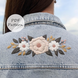 以下が含まれることがあります： 刺繍が施されたライトブルーのデニムジャケット。刺繍は、柔らかいピンク色の大きな中央の花、白い花、灰色の葉、金とピンクのアクセントが特徴です。「PDF Pattern」というテキストが白い円の中にあります。