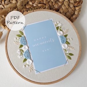 May include: A light blue card with the words "HAPPY moments" and "4 x 6" is framed by a wooden embroidery hoop. The embroidery features blue, white, and green floral designs. The text "PDF Pattern" is in a white circle.