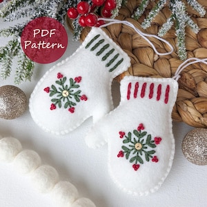 May include: Two white felt mitten ornaments with snowflake embroidery. The mittens have green and red accents, with a gold sequin center. The text "PDF Pattern" is in a red circle in the upper left corner. White string loops are attached for hanging.
