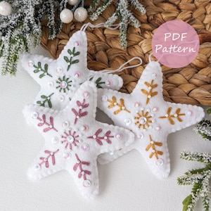 May include: Three white felt star decorations with embroidered floral patterns in green, gold, and pink, embellished with sequins. Each star has a hanging loop. The image also includes a pink circle with the text "PDF Pattern".