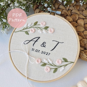 Op de afbeelding: Geborduurde borduurring met de letters "A & T" en de datum "11.07.2027" in zwart garen. Het ontwerp bevat roze rozen en witte bloemen met groene bladeren. De tekst "PDF Pattern" staat in een roze cirkel.