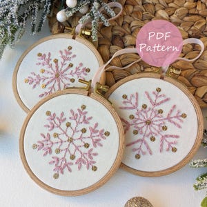 Op de afbeelding: Drie geborduurde sneeuwvlokornamenten. Elk ornament heeft een roze sneeuwvlokontwerp met gouden accenten op een witte stof, gezet in een houten ring. De tekst "PDF Pattern" is zichtbaar. Lintjes zijn bevestigd om op te hangen.