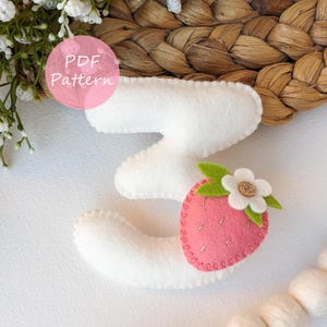 Peut inclure: Chiffre trois en feutre blanc avec un accent de fraise rose et une fleur blanche avec un centre doré. La fraise a des feuilles vertes et est cousue avec du fil doré. Le texte "PDF Pattern" est dans un cercle rose.