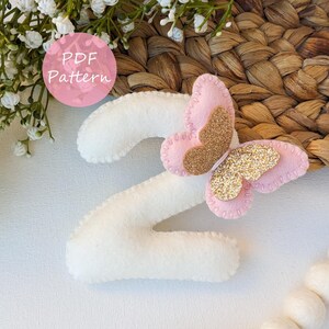Peut inclure: Un chiffre deux en feutre blanc, orné d'un papillon rose aux ailes dorées pailletées. Le papillon est cousu sur le chiffre. L'image comprend également le texte "PDF Pattern" dans un cercle rose.