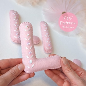 May include: Pink felt letter "L" with white stitching and sequin accents, held by hands. The letter is part of a set, as indicated by the "PDF Pattern" text in a pink circle. Additional felt pieces are visible in the background.