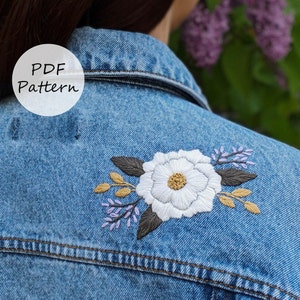 Puede incluir: Una chaqueta vaquera con una flor blanca y hojas verdes bordadas en la espalda. El bordado es de estilo floral con acentos morados y amarillos. El texto "PDF Pattern" es visible en un círculo blanco en el lado izquierdo de la imagen.
