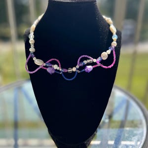 May include: A handcrafted necklace featuring a mix of white, purple, pink, and blue beads. The necklace is displayed on a black velvet bust. The design incorporates various bead shapes and sizes, creating a unique and colorful accessory.