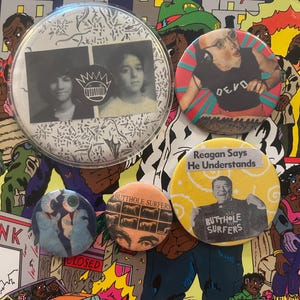 Puede incluir: Una colección de botones redondos con varios diseños. Un botón presenta a dos personas en una foto en blanco y negro. Otro muestra a una persona con auriculares y la palabra "DEVO". Otros botones tienen el texto "Butthole Surfers" y una foto de Ronald Reagan.