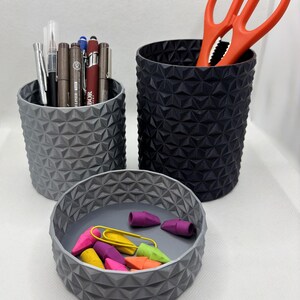 May include: Three geometric patterned desk organizers in gray and black. One tall gray container holds pens, another black container holds red scissors, and a shallow gray dish contains erasers and a yellow paperclip. The containers are cylindrical.