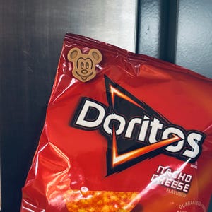 May include: A red bag of Doritos Nacho Cheese flavored tortilla chips with a Mickey Mouse-shaped wooden magnet attached. The bag has a black and white logo and a picture of the chips. The magnet is light brown.