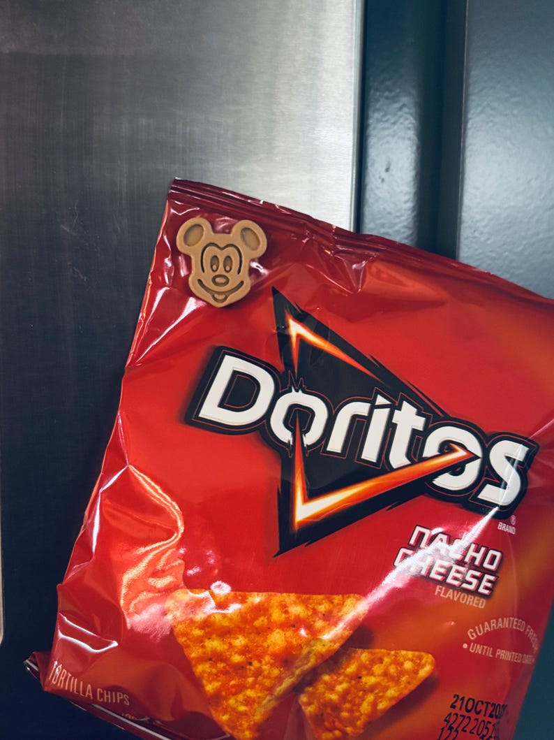 May include: A red bag of Doritos Nacho Cheese flavored tortilla chips with a Mickey Mouse-shaped wooden magnet attached. The bag has a black and white logo and a picture of the chips. The magnet is light brown.