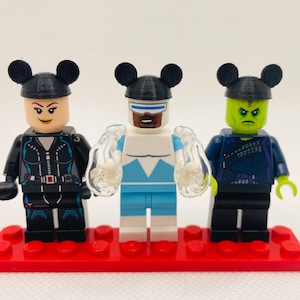 May include: Three Lego minifigures wearing black Mickey Mouse ears. The minifigures are standing on a red Lego baseplate. The minifigure on the left is wearing a black outfit with a white belt. The minifigure in the middle is wearing a white and blue outfit. The minifigure on the right is wearing a green and black outfit.