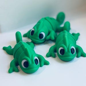 May include: Three bright green chameleon figurines with large, expressive eyes. The chameleons are lying down, with their legs and tails extended. The figurines have a textured, 3D-printed appearance, suggesting a playful, decorative item.