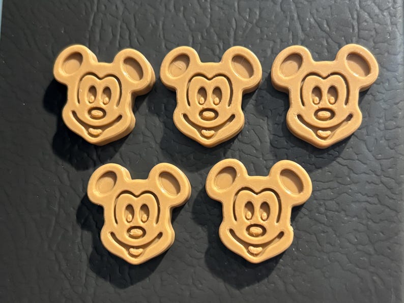 May include: Five tan-colored Mickey Mouse-shaped embellishments. Each has a smiling face, two large ears, and a detailed facial outline. The embellishments are arranged on a dark surface.