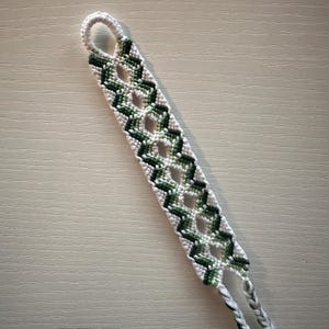 May include: A handmade woven bracelet featuring a repeating geometric zigzag pattern in shades of dark green, light green, and white. The design includes open diamond-shaped gaps throughout the center, ending in a circular loop and two braided ties.