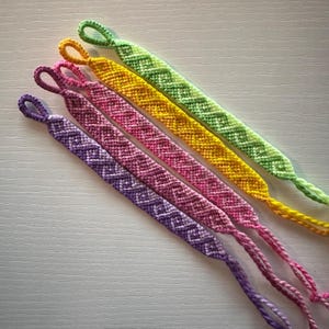 Könnte beinhalten: Fünf Freundschaftsbänder in einem Regenbogenfarbverlauf. Die Armbänder sind mit einem Chevron-Muster gefertigt und am Ende mit einem Knoten gebunden.