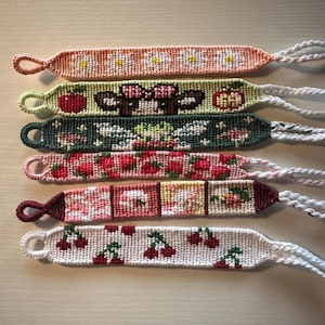 May include: Six colorful friendship bracelets with various patterns, including daisies, apples, a cartoon animal, strawberries, and cherries. The bracelets are made with colorful embroidery floss and have white tassels.