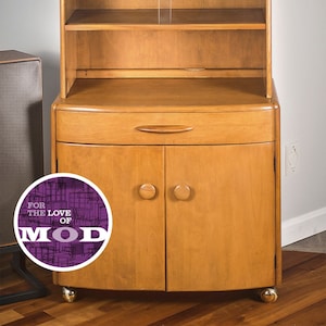 May include: A mid-century modern hutch with a glass-front cabinet and two doors. The hutch is made of wood and has a light brown finish. The cabinet has two shelves and the doors have round knobs. The hutch is on wheels and has a drawer below the cabinet.