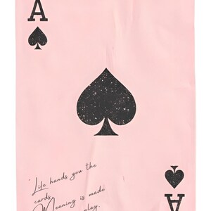 Puede incluir: Una carta de juego rosa con el As de Picas. La carta tiene un gran símbolo de pica negro en el centro, con símbolos de pica más pequeños y la letra "A" en las esquinas. También tiene el texto "Life hands you the cards, Meaning is made by how you play."